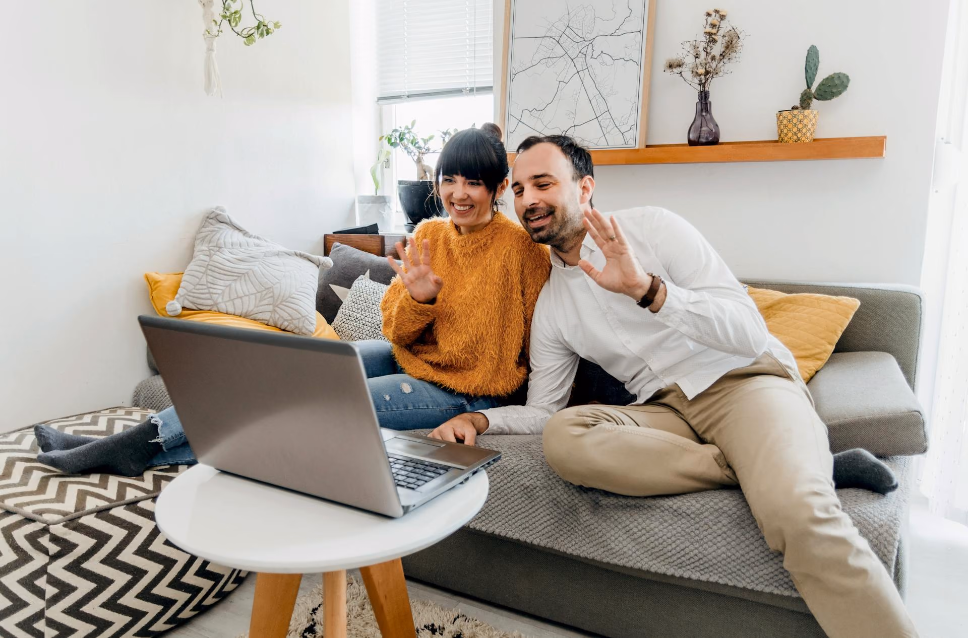 A couple sitting on a living room floor, waving at a laptop during a virtual meeting