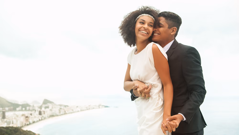 A happy couple in wedding attire embracing on a beach with a coastal town in the background