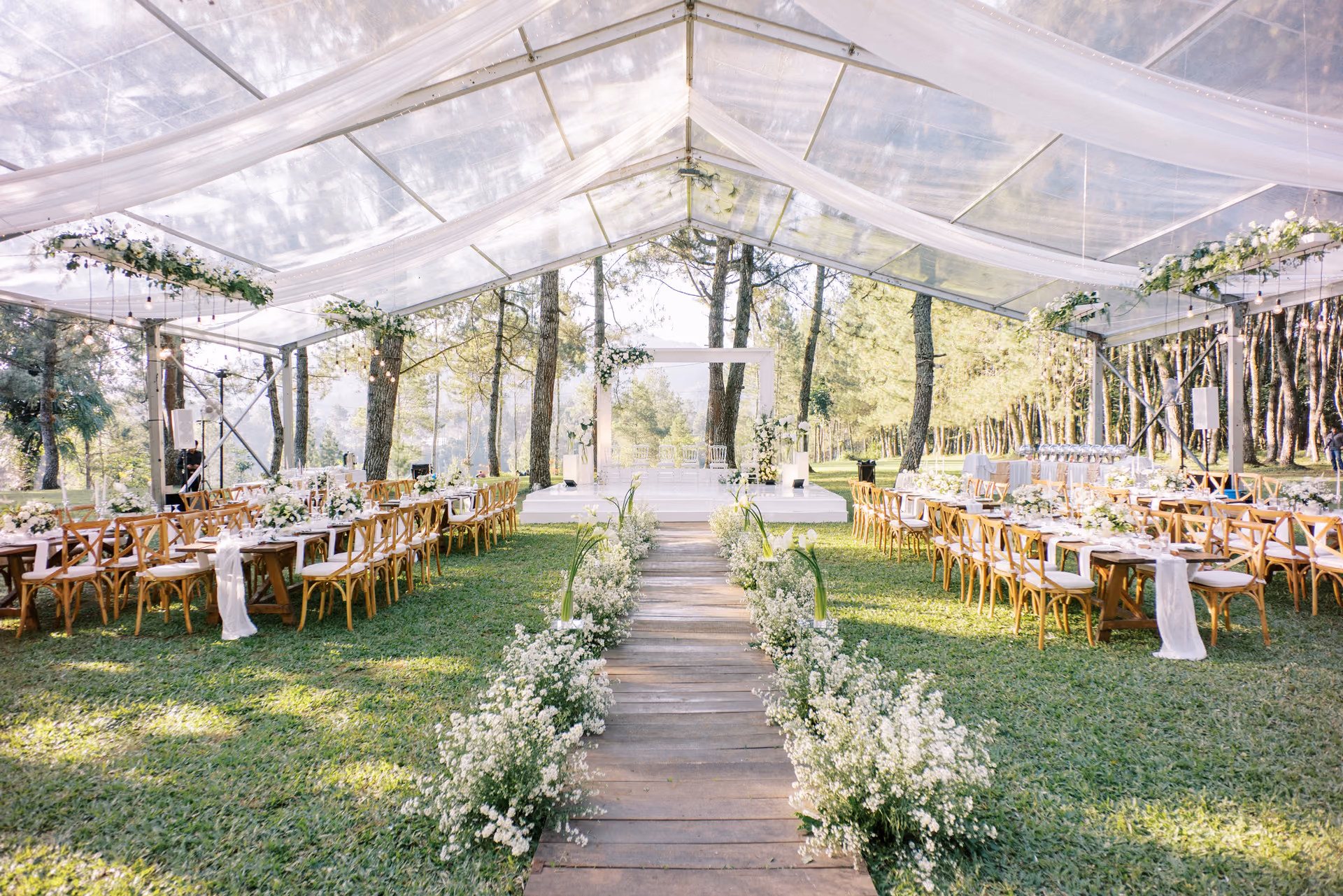 Long reception tables set up on a wooden walkway under a sheer, draped tent outdoors